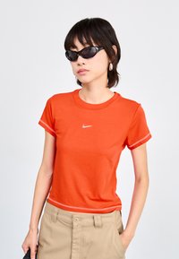 Narancssárga, rövid ujjú póló kerek nyakkal, mellén egy kis fehér Nike logóval. Bézs nadrággal párosítva.
