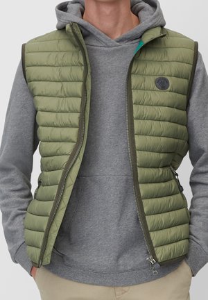 Persoon draagt een groene doorgestikte bodywarmer over een grijs hoodiesweater, handen in de zakken, beige broek zichtbaar aan de onderkant.