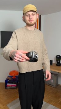 Schwarzer Ledergürtel mit einer silbernen rechteckigen Schnalle, gehalten von einer Person, die einen beigen Pullover und schwarze Hose trägt, in einem Raum mit hellem Holzboden.