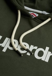 Felpa verde con scritta bianca crepata "superdry", caratterizzata da lacci color off-white con punte in metallo. È visibile una texture di tessuto morbido.