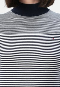 Mujer vistiendo un suéter de cuello alto a rayas blancas y negras con un pequeño logo rectangular rojo, blanco y azul en el pecho.