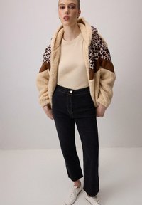 Chaqueta de piel sintética beige acogedora con mangas de estampado de leopardo, detalles en marrón y capucha, combinada con jeans negros de cintura alta y zapatillas blancas.