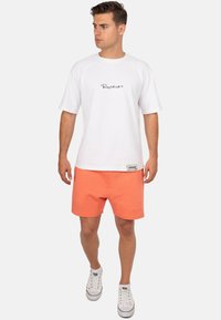Witte t-shirt met korte mouwen en zwart scriptlogo, gecombineerd met koraalkleurige shorts en witte sneakers. Casual, relaxte pasvorm met ademende stof.