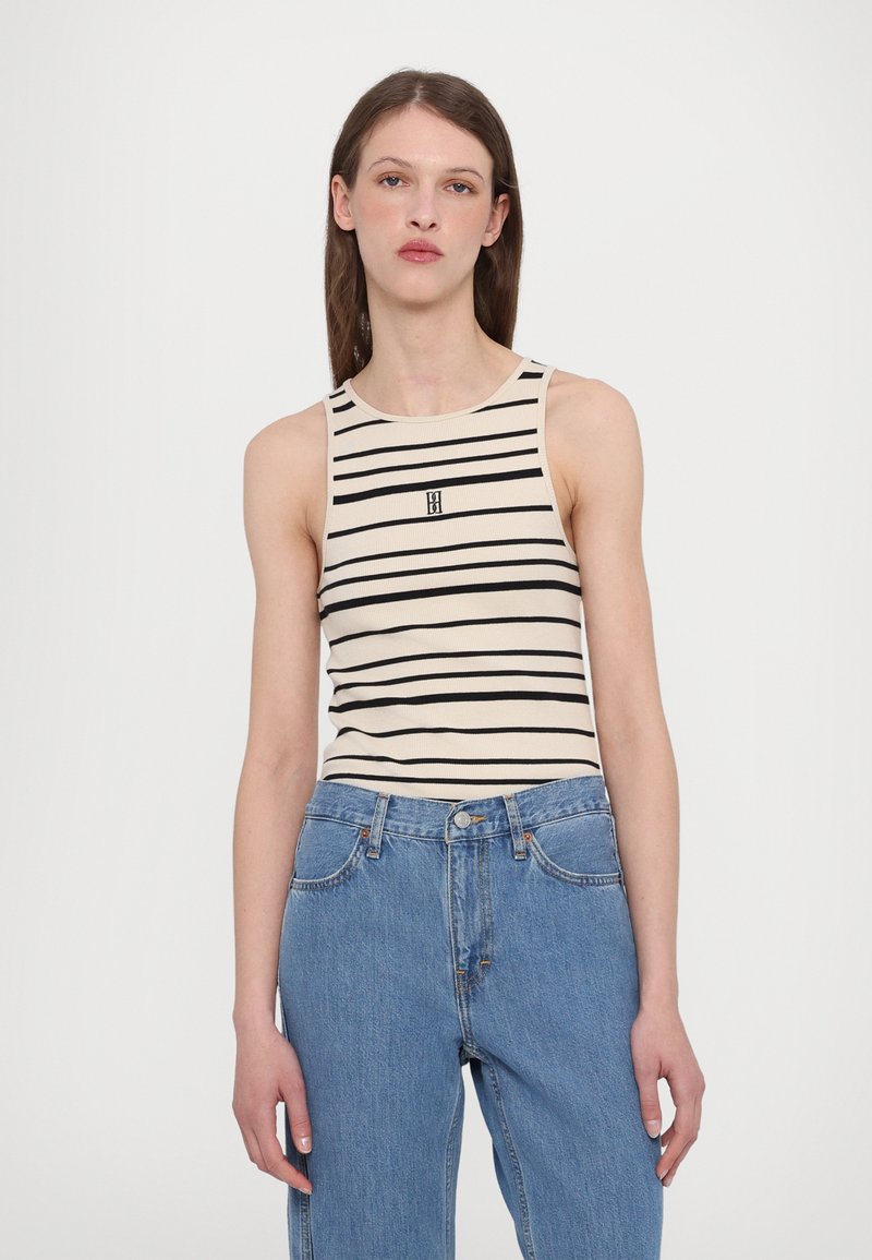 Gestreiftes ärmelloses Oberteil in Creme und Schwarz, gerippte Textur, runder Ausschnitt, mit kleinem Logo in der Mitte. Kombiniert mit lockeren blauen Jeans.