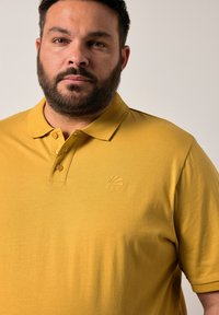 Polo in giallo senape realizzato in tessuto morbido. Presenta un colletto classico, una patta con tre bottoni e un logo ad ocra del sole in rilievo sul petto.
