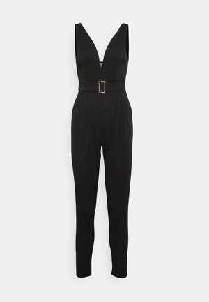 Zwarte mouwloze jumpsuit met diepe V-hals, getailleerde tailleband met gesp en taps toelopende pijpen tegen een effen achtergrond.