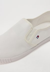 Fehér vászon slip-on cipő, gumitalppal, piros, fehér és kék hímzett logóval. Sima textúrájú és letisztult dizájnnal rendelkezik.
