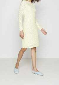 Kvinna som bär en knälång, långärmad klänning i krämfärgad spets och ljusblå flätade ballerinas, stående framför en enfärgad vit bakgrund.