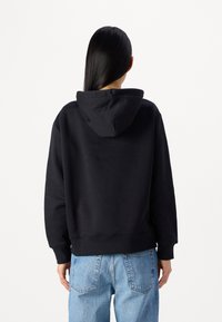 Felpa nera con cappuccio, dal tessuto morbido, con polsini e orlo a coste, silhouette rettangolare e dettagli di cucitura visibili, abbinata a jeans blu chiaro.