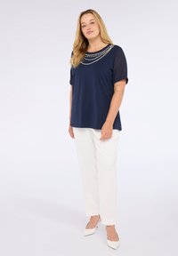 Mujer con blusa azul marino con escote de cuentas, pantalones blancos y tacones blancos, de pie contra un fondo claro y liso.
