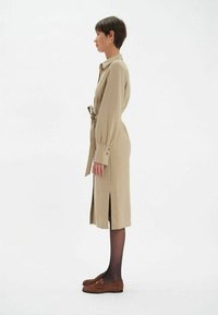 Robe chemise beige avec une ceinture à la taille, manches longues, fentes latérales et poignets boutonnés, associée à des mocassins bruns et des collants noirs.