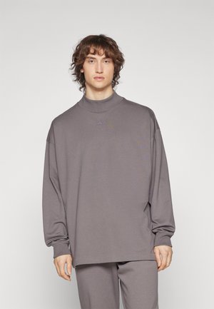 Oversized grey long-sleeve top with a high collar, featuring wide sleeves and smooth texture. Includes subtle branding on the chest.
