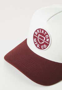 Cappellino bianco e bordeaux con visiera curvata, che presenta un logo ricamato circolare con la scritta "Brixton MFG Co." sul davanti.
