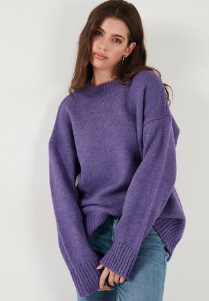 Purple knitted sweater with a crew neck, loose fit, and ribbed cuffs. Features side slits and a textured finish. Paired with light jeans.
