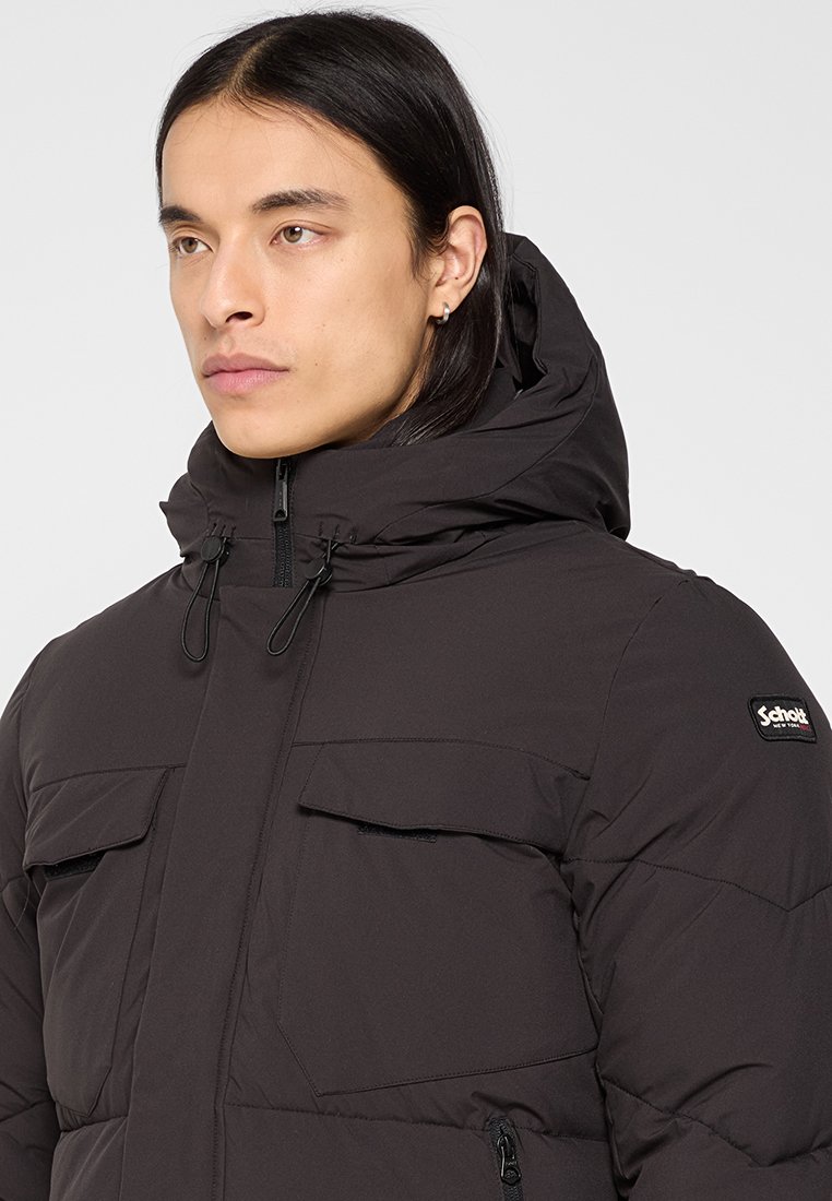 Jeune personne avec de longs cheveux foncés portant une veste rembourrée noire avec capuche et poches avant, tournée vers la gauche sur un fond clair.