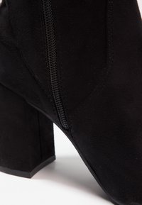 Bottine en daim noir avec talon bloc et fermeture éclair latérale sur fond blanc.