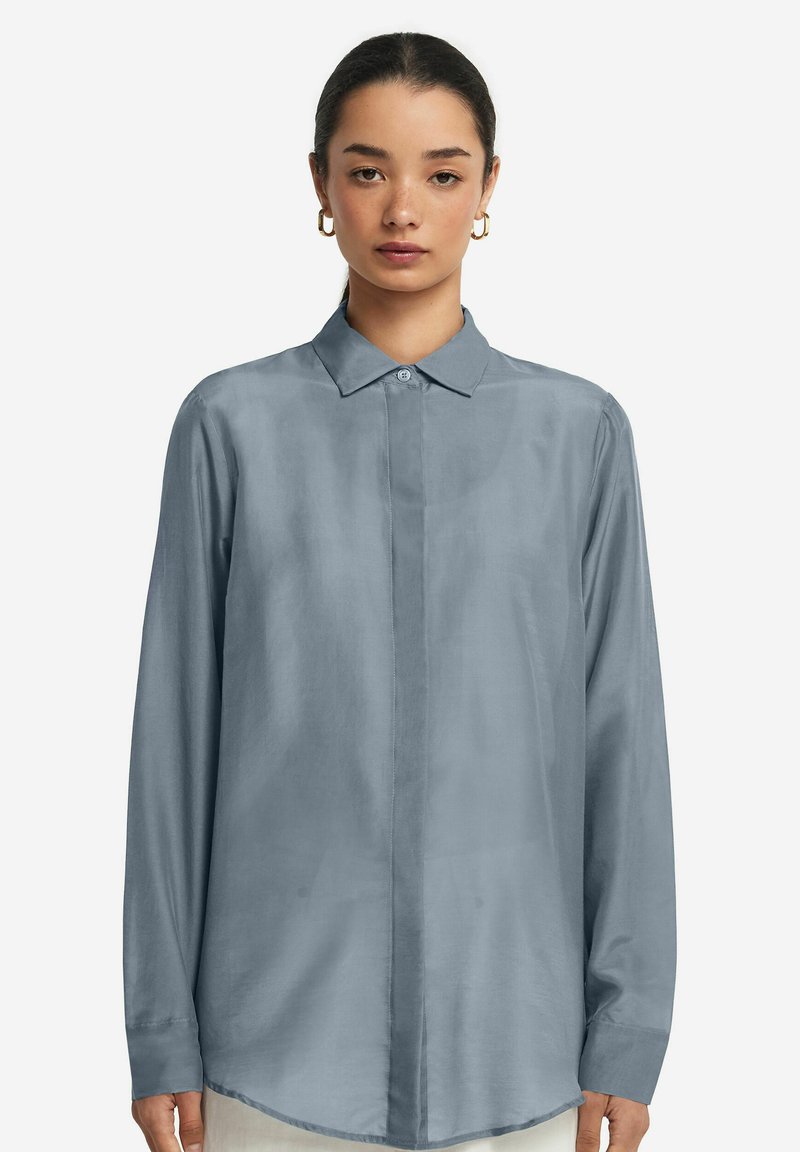 Chemise bleu clair à manches longues avec un col classique, une fermeture à boutons et une texture lisse. Présente un ourlet arrondi et un design simple.