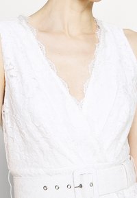 Robe en dentelle blanche avec un décolleté en V profond, présentant des bords en dentelle effilochés et une large ceinture en tissu avec des accents en argent. Texture élégante à motif floral.