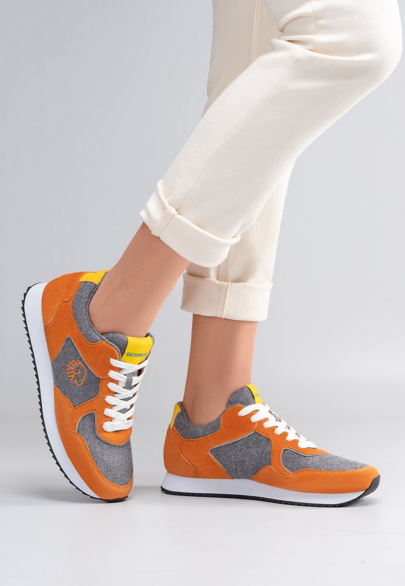 Benen in opgerolde crèmekleurige broek met oranje, grijze en gele sneakers met witte veters en zwarte rubberen zolen op een lichtgrijze achtergrond.