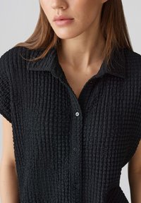 Camicia collared nera, di tessuto testurizzato, con maniche corte, caratterizzata da una chiusura a bottoni e un motivo di tessuto sgualcito per un dettaglio aggiuntivo.