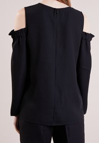 Svart topp med kall axel-design, volangärmar och en dragkedja bak. Släta tyg med avslappnad passform.