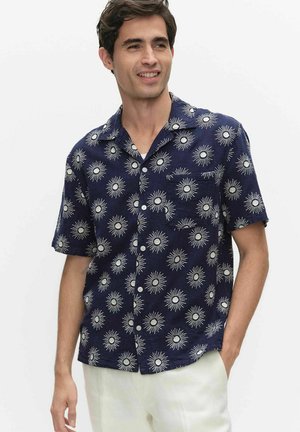 Mężczyzna w granatowej koszuli z krótkim rękawem zapinanej na guziki, zdobionej białymi wzorami przypominającymi promienie słońca, i jasnych spodniach, uśmiechający się i patrzący w bok.