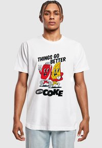T-shirt blanc à manches courtes avec un design de dessin animé représentant une cannette de soda rouge et des frites jaunes, le texte indique "LES CHOSES S'AMÉLIORENT AVEC DU COKE."
