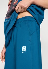 Persoană care ridică tricoul albastru pentru a arăta talia elastică a pantalonilor scurți albaștri Nike cu litera „S” albă și logo-ul swoosh pe coapsă.
