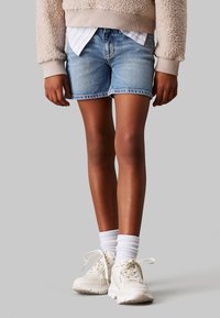 Bermuda in denim azzurro chiaro con orlo sfrangiato. Abbinata a un maglione beige dalla texture particolare e una camicia bianca con colletto, completata da sneakers bianche.