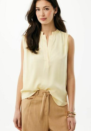 Frau mit langem, dunklem Haar, die eine ärmellose cremefarbene Bluse und beigefarbene, hoch taillierte Hosen mit gebundenem Gürtel trägt, steht vor einem weißen Hintergrund.