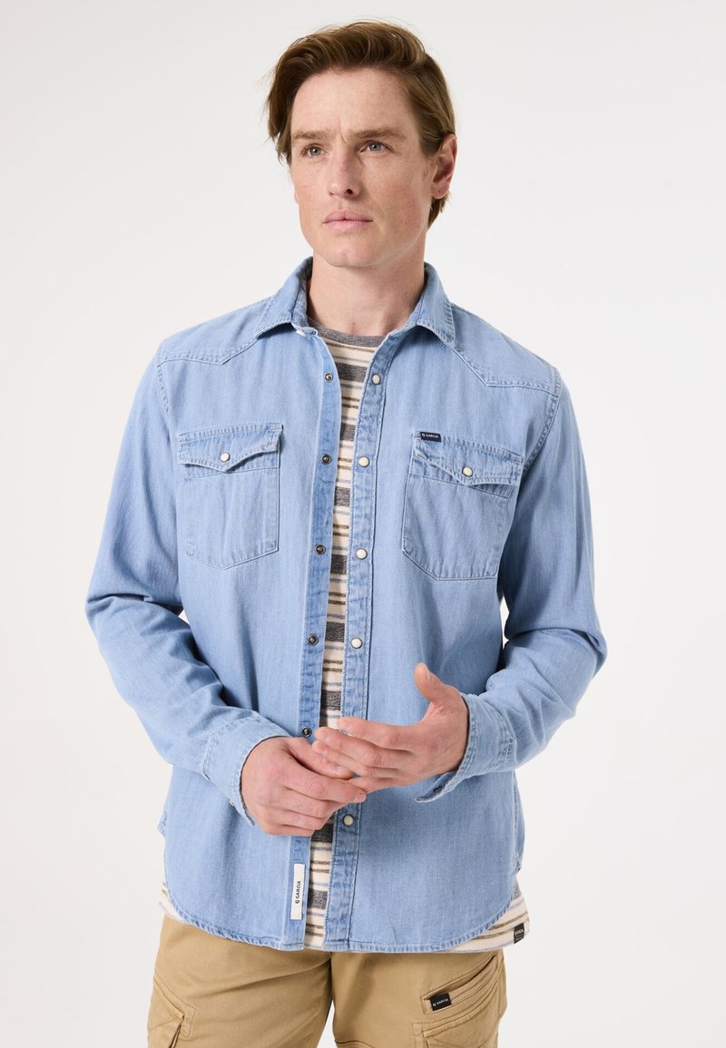 Lichtblauwe denim shirt met lange mouwen, knoopsluiting aan de voorkant, twee borstzakken en een gebogen zoom, gedragen over een gestreept T-shirt.