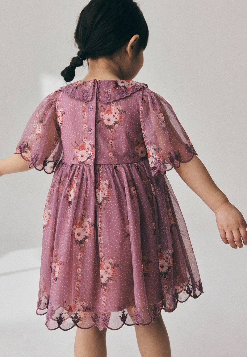 Jeune enfant aux cheveux foncés tressés, portant une robe violette à fleurs avec des manches transparentes et des bords festonnés, debout les bras écartés.