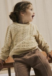 Maglione a maglia beige chiaro con un motivo testurizzato, abbinato a pantaloni marroni con puntini in rilievo. Bambino appoggiato a una sedia.