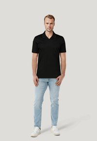 Czarna koszulka polo z krótkim rękawem, płaskim kołnierzykiem i gładkiego materiału. W połączeniu z jasnoniebieskimi dżinsami i białymi tenisówkami. Neutralne tło.