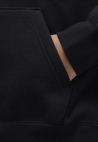 Close-up do tecido de um sweatshirt preto com um bolso canguru, punhos canelados e uma textura suave e macia. Mão parcialmente visível no bolso.