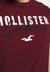 Mężczyzna nosi bordową koszulkę Hollister z haftem "Hollister California" oraz białym logo ptaka na piersi.
