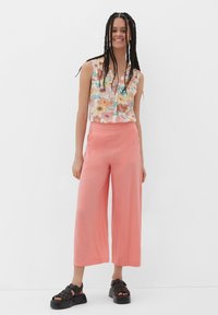 Jonge vrouw met gevlochten haar, die een mouwloze bloeiende blouse, koraalkleurige wijde broek en zwarte plateau sandalen draagt, staand en lachend.