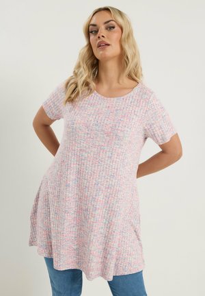 Blonde Frau mit welligem Haar trägt eine kurzärmelige, pastellfarbene, gerippte Tunika über blauen Jeans und hat die Hände hinter dem Rücken verschränkt.