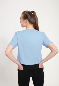 Světlemodré bavlněné tričko s krátkým rukávem, kulatým výstřihem a volným střihem, ve spojení s černými džíny. Model stojí se zkříženýma rukama v kapsách.