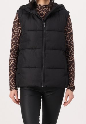 Zwart doorgestikte mouwloze capuchonvest gedragen over een lange mouw bruine en zwarte luipaardprint top en zwarte broek.