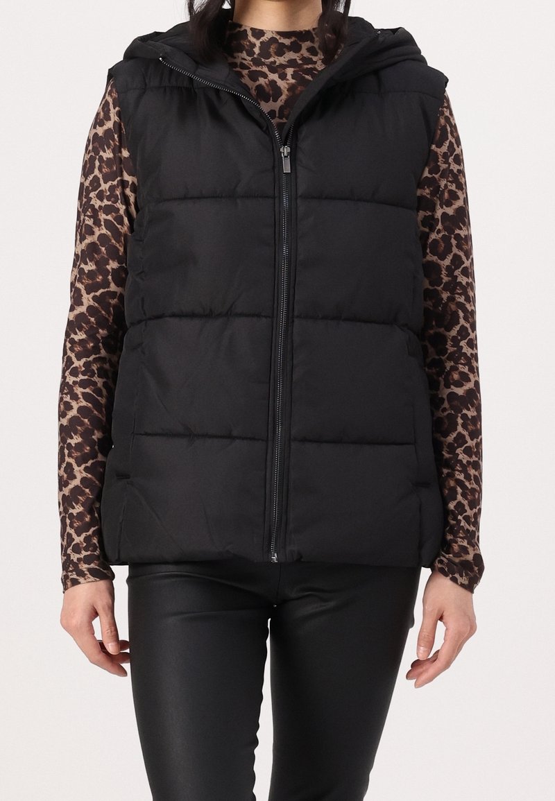 Veste sans manches matelassée noire à capuche portée sur un haut à manches longues imprimé léopard marron et noir et un pantalon noir.