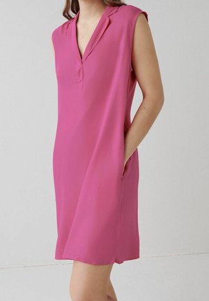 Mujer con un vestido sin mangas, de longitud hasta la rodilla, de color rosa brillante, con escote en V y manos en los bolsillos laterales, sobre un fondo liso.