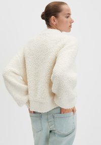 Maglione color crema con una maglia texturizzata e soffice, maniche ampie e orlo aderente, abbinato a jeans blu chiaro con tasche posteriori.