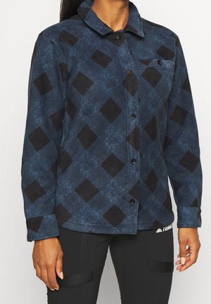 Frau trägt ein blaues Hemd mit schwarzen Rautenmuster und schwarze Hose mit strukturierten Einsätzen an den Oberschenkeln.