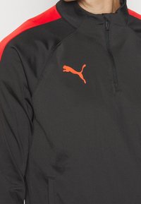 Schwarze Sportjacke mit roten Akzenten, Halb-Reißverschlusskragen und einem dezenten orangen Puma-Logo auf der Brust. Glattes Polyestergewebe.