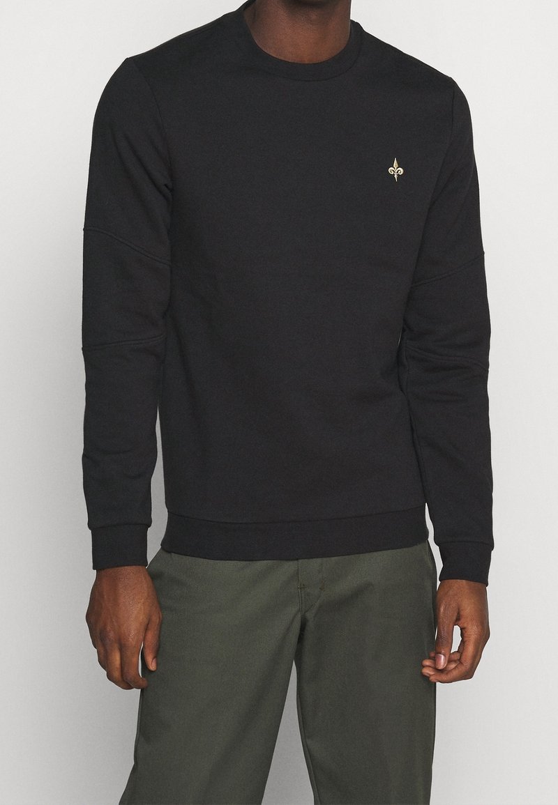 Homme portant un sweat-shirt noir à manches longues avec un petit emblème doré fleur-de-lis sur la poitrine et un pantalon vert olive.