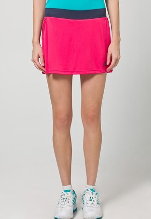 Jupe de sport rose avec une taille noire, fabriquée en tissu léger. Caractérisée par un ourlet plat et associée à des chaussures de sport blanches et teal.