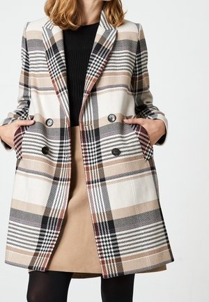 Donna che indossa un cappotto doppiopetto a quadri beige, nero e bianco sopra una maglietta nera e una gonna cammello con le mani nelle tasche.
