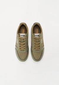 Sneaker verde oliva con tomaia in tessuto testurizzato, lacci piatti e suola in gomma bianca. Presenta un'etichetta con logo marrone sulla linguetta.