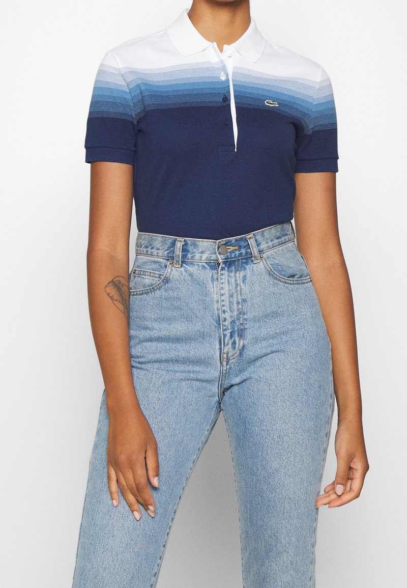 Personne portant un polo à manches courtes avec des rayures dégradées bleues et un jean en denim bleu clair taille haute, montrant un tatouage sur le bras gauche.